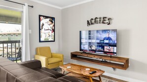 A living room with a brown leather sofa, a yellow armchair, a wooden coffee table, and a mounted TV displaying various channels. - The Zone: Blocks from Texas A&M, Comfy King Bed (Bryan)