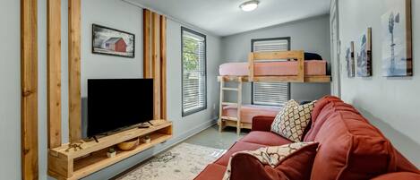 Una sala de estar moderna con una televisión sobre un mueble de madera, un sofá rojo, una cama litera y un ventanal con persianas.