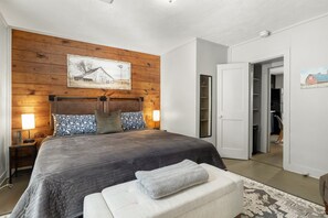 A bedroom with a wooden headboard, a large bed with a dark quilt, two bedside lamps, and a painting on the wall.