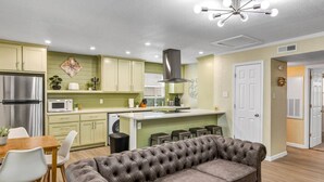 A modern kitchen with green cabinets, a stainless steel refrigerator, and a central island. There is a dining area with a brown leather sofa and a table.