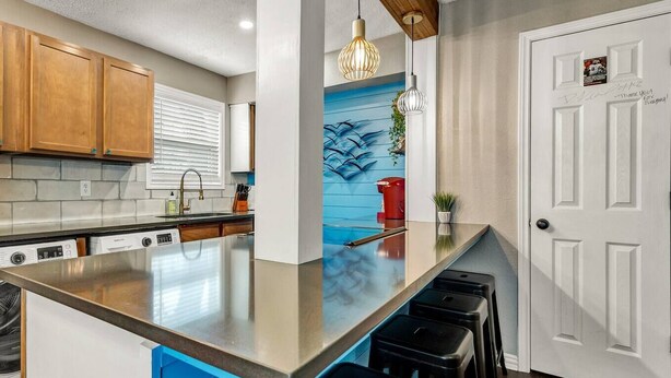 A modern kitchen with a large island, wooden cabinets, and a blue accent wall. - Kelly's Cove: 1st Flr 2 Bdrm-Walk to A&M-King Beds (Bryan)