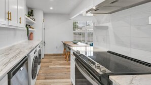 Moderni keittiö, jossa on valkoiset kaapit, sisäänrakennettu uuni, liesi sekä ruokailutila pöytineen ja tuoleineen.