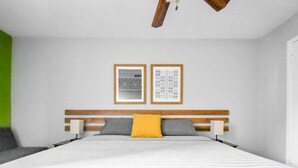 Une chambre moderne avec une tête de lit en bois, un grand lit recouvert d’une couette à rayures blanches et grises, deux oreillers (dont un jaune) et deux tableaux encadrés accrochés au mur.