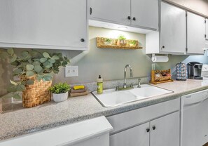 Una cocina con armarios blancos, encimera de granito, un fregadero y una estantería con plantas.