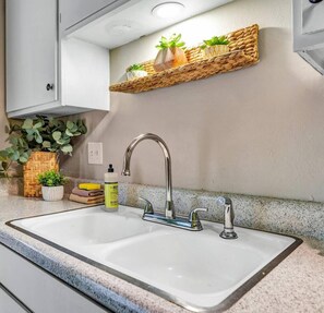 Una cocina con un lavamanos doble, un grifo de acero inoxidable y un estante tejido sobre el lavamanos con plantas en macetas.