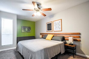 Un dormitorio con cama, mesita de noche, lámpara, ventilador de techo y dos cuadros en la pared.
