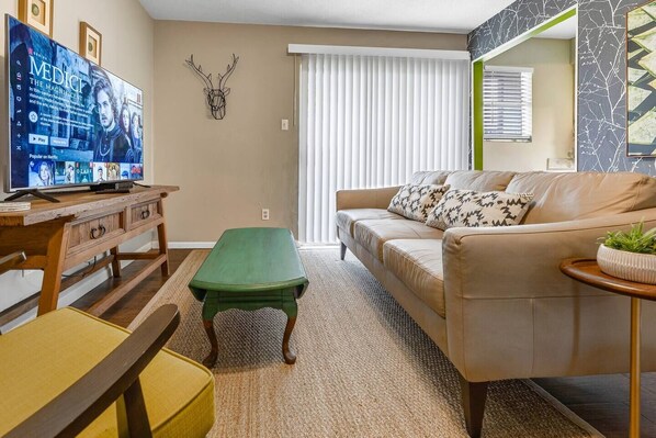 Una sala de estar con televisor, una mesa de centro verde, un sofá color beige con cojines estampados, una mesita de noche de madera y una decoración de cabeza de ciervo montada en la pared.