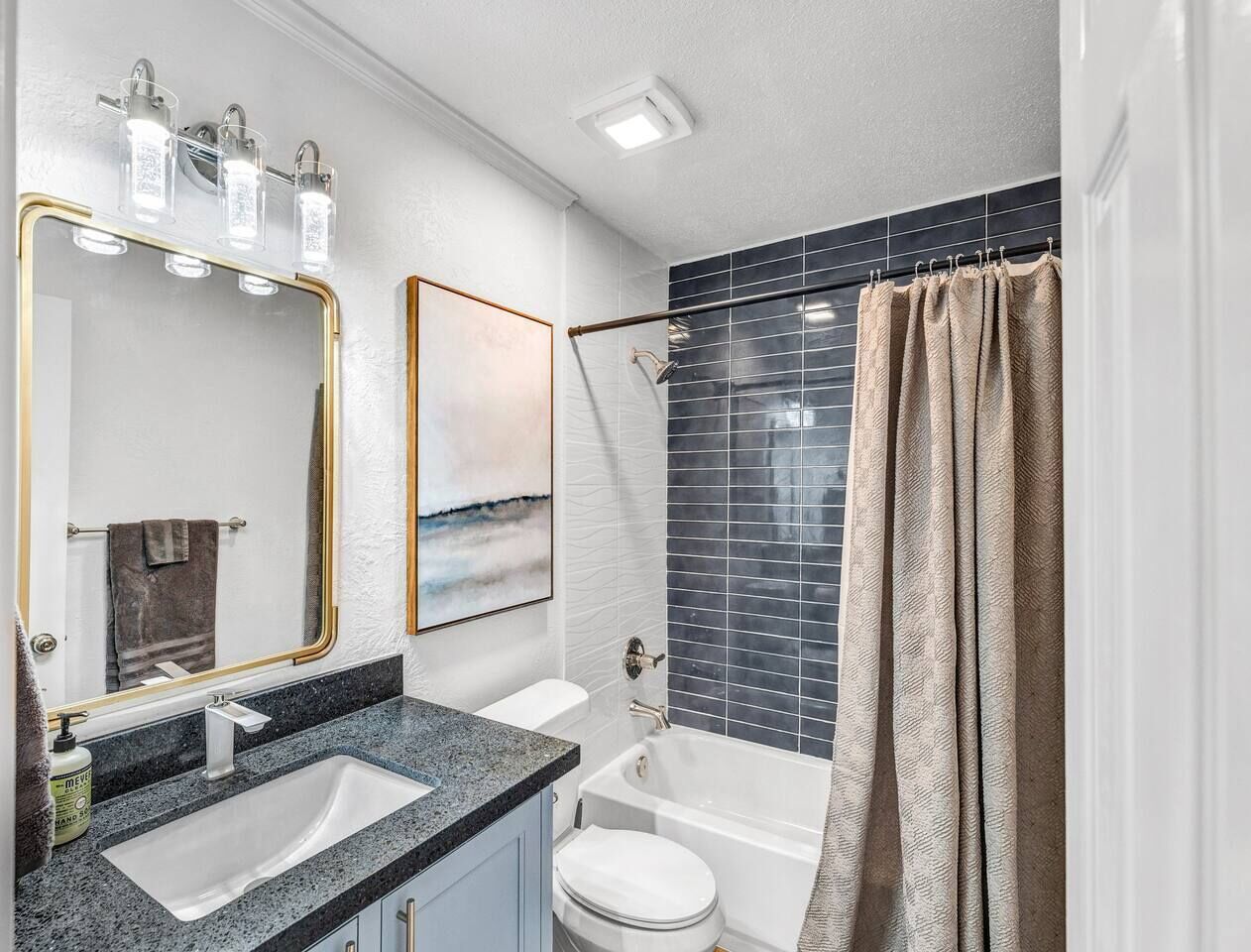 Un baño moderno con encimera de granito, un lavabo blanco, un espejo con marco dorado, una ducha con azulejos oscuros y una cortina de ducha color beige.
