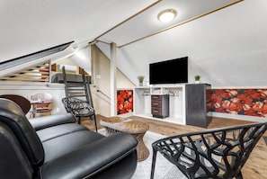 A modern loft-style living space with a black leather sofa, a wicker coffee table, and a black metal chair. A flat-screen TV is mounted on the wall, and there is a staircase leading to an upper level.