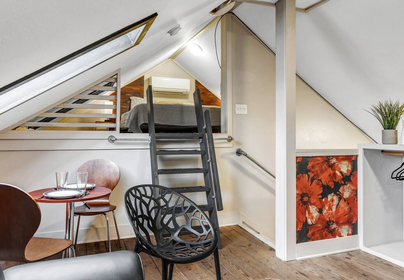 A loft bedroom with a sloped ceiling, a wooden floor, a ladder, a dining table with chairs, and a painting.