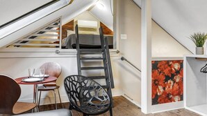 A loft bedroom with a sloped ceiling, a wooden floor, a ladder, a dining table with chairs, and a painting. - Poppy: Quirky, Walk to A&M, Comfy King Bed, Fun Space (Bryan)