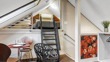 A loft bedroom with a sloped ceiling, a wooden floor, a ladder, a dining table with chairs, and a painting.