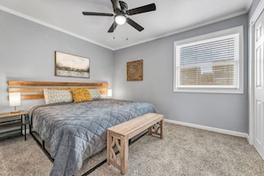 Une chambre à coucher avec un lit, une tête de lit en bois, un ventilateur de plafond, une fenêtre avec des stores et un sol recouvert de moquette.