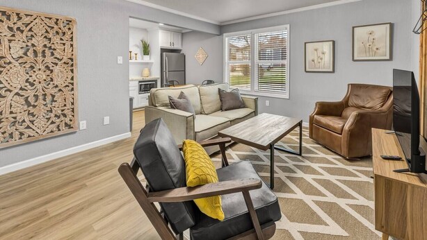 A modern living room with a sofa, a leather armchair, a coffee table, and a TV. There is a kitchen in the background with white cabinets and a refrigerator. - Rowan: 1st Floor, King Beds/Close to A&M (Bryan)
