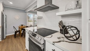 A modern kitchen with a gas stove, oven, and a built-in range hood. There is a dining area with a table and chairs, and a window providing natural light. - Rowan: 1st Floor, King Beds/Close to A&M (Bryan)