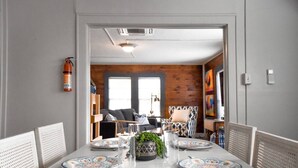 Un coin repas avec une table ronde dressée pour quatre personnes, un extincteur accroché au mur, et une vue sur un salon avec un canapé et un téléviseur.