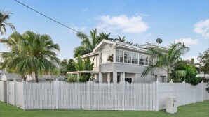 A two-story house with a white picket fence, surrounded by palm trees and a clear sky.