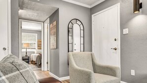 A hallway with a checkered armchair, a door, and a mirror on the wall. - The 12: 1st Floor 2 Bdrm, Walk to A&M, King Beds (Bryan)