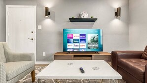 A living room with a flat-screen TV mounted on a wooden stand, a white coffee table, a patterned armchair, and a brown leather sofa. - The 12: 1st Floor 2 Bdrm, Walk to A&M, King Beds (Bryan)