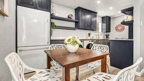 A modern kitchen with dark cabinets, a white refrigerator, and a wooden dining table with white chairs. - The 12: 1st Floor 2 Bdrm, Walk to A&M, King Beds (Bryan)