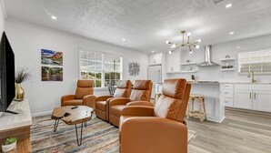 A modern living room with leather sofas, a coffee table, and a TV. The room has a large window, a kitchen area with white cabinets, and a ceiling light fixture. - The Dominion: 2 Bdrm/Walk to A&M/Comfy King Beds (Bryan)