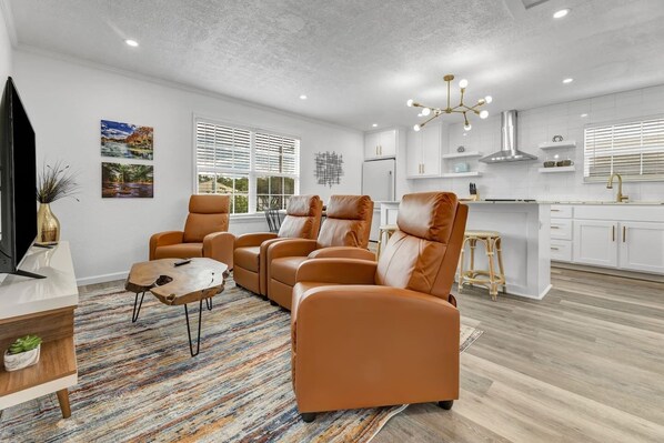 A modern living room with leather sofas, a coffee table, and a TV. The room has a large window, a kitchen area with white cabinets, and a ceiling light fixture. - The Dominion: 2 Bdrm/Walk to A&M/Comfy King Beds (Bryan)