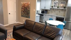 A living room with a brown leather sofa, a wooden coffee table, and a dining area with a white table and blue chairs. - Wildflower: Very Near Texas A&M-Comfy King Beds (Bryan)