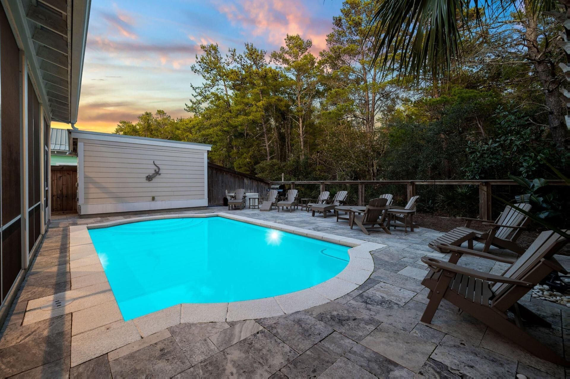 A backyard with a swimming pool, surrounded by stone tiles, and furnished with wooden chairs and a grill.