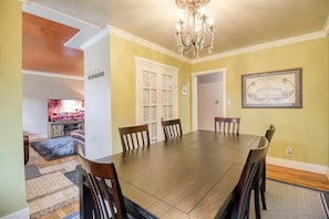 A dining room with a large wooden table and six chairs, a chandelier, and a framed picture on the wall.