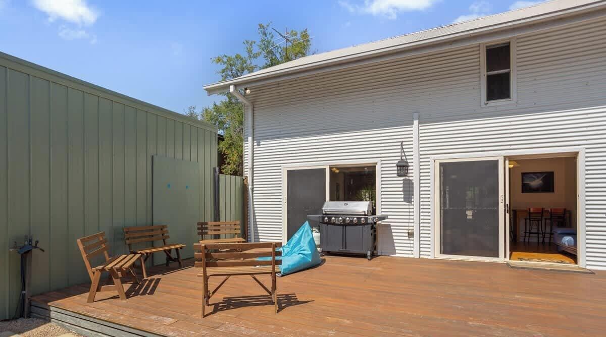 Une terrasse avec des meubles en bois et un barbecue, un abri de jardin vert et un bâtiment blanc avec une porte de garage.