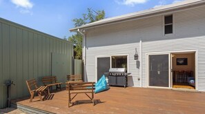 A patio with wooden furniture and a grill, a green shed, and a white building with a garage door.