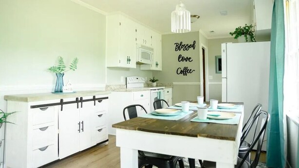 A compact kitchen with white cabinets, a wooden dining table set for four, and a sign that reads "Blessed love Coffee". - Peaceful 3BR/2BA House Near EVERYTHING w/WIFI  (Dothan)