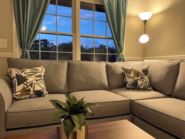 A sectional sofa with patterned cushions, a potted plant, and a floor lamp in a room with a window and curtains. - Peaceful 3BR/2BA House Near EVERYTHING w/WIFI (Dothan)