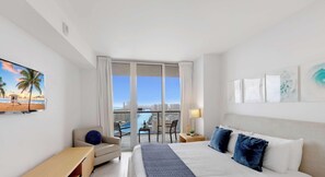 A hotel room with a bed, a chair, a desk, a TV, and a balcony with a city view.