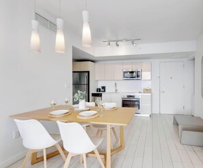 Une cuisine moderne dotée d’appareils électroménagers en acier inoxydable, un coin repas avec une table en bois et des chaises blanches, et un éclairage suspendu.