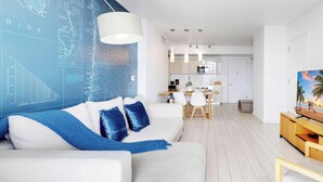 Ein modernes Wohnzimmer mit einem weißen Sofa, blauen Kissen, einem Essbereich mit weißem Tisch und Stühlen, einer Küche mit weißen Schränken und einem Fernseher auf einem Holzschrank.