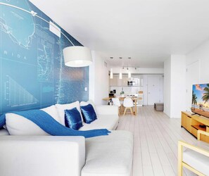 A modern living room with a white sofa, blue cushions, a dining area with a white table and chairs, a kitchen with white cabinets, and a TV on a wooden stand.