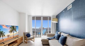A modern living room with a large window, a sofa, a chair, a TV, and a balcony with a view of the city. - Upper Apartment + Pool + Gym + Amazing view (Hallandale Beach)