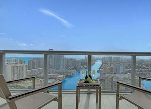 A balcony with a table, two chairs, and a view of a cityscape and water body. - Upper Apartment + Pool + Gym + Amazing view (Hallandale Beach)
