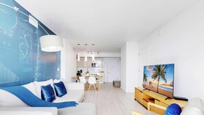 Uma sala de estar moderna com um sofá branco, almofadas azuis, uma área de jantar com uma mesa e cadeiras brancas, uma cozinha com armários brancos e uma TV sobre um suporte de madeira.