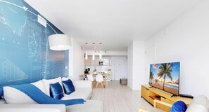 A modern living room with a white sofa, blue cushions, a dining area with a white table and chairs, a kitchen with white cabinets, and a TV on a wooden stand. - Upper Apartment + Pool + Gym + Amazing view (Hallandale Beach)