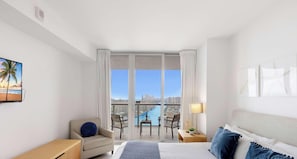 A hotel room with a bed, a chair, a desk, a lamp, and a view of a cityscape through large windows.