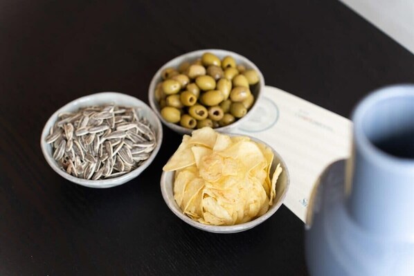 Three bowls containing sunflower seeds, green olives, and potato chips on a dark surface.