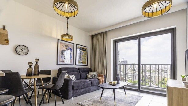 Una sala de estar moderna con un sofá gris, una mesa de comedor y sillas. Hay dos lámparas colgantes, un ventanal con vistas, y un reloj en la pared.