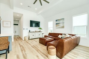 A modern living room with a brown leather sofa, a dining area with a white table and wicker chairs, and a large flat-screen TV mounted on the wall.