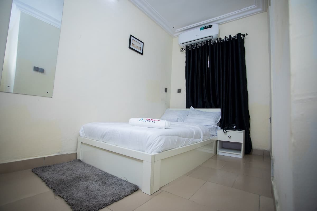A neatly arranged bedroom with a bed, a bedside table, a nightstand, a small rug, a wall-mounted air conditioner, and a framed picture.