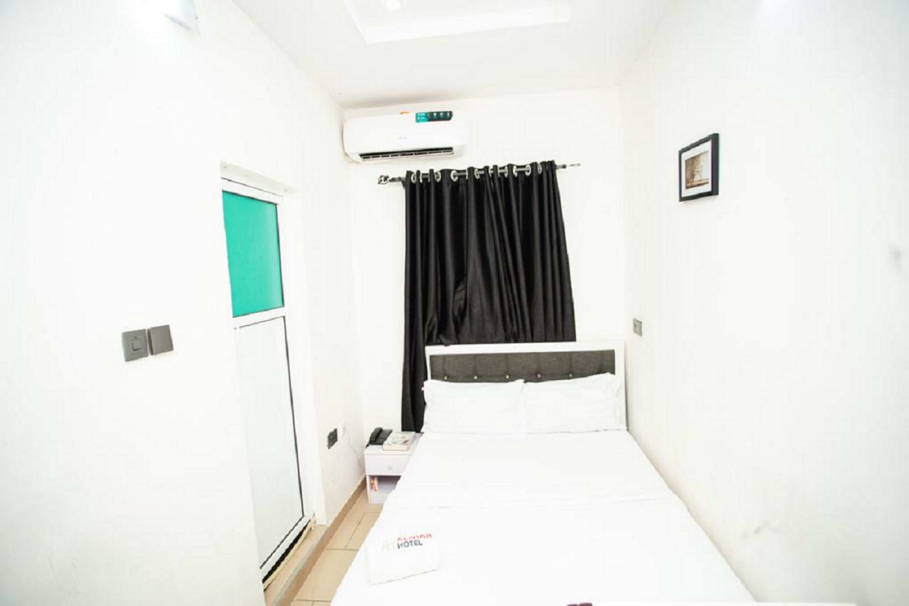 A hotel room with a bed, an air conditioning unit, a curtain, a bedside table, and a nightstand.