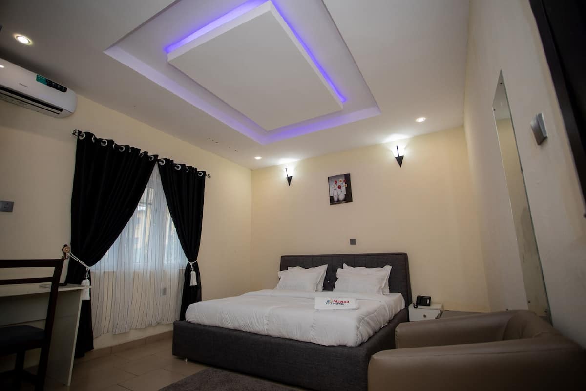 A hotel room with a bed, a desk, a chair, a sofa, an air conditioning unit, and a ceiling light.