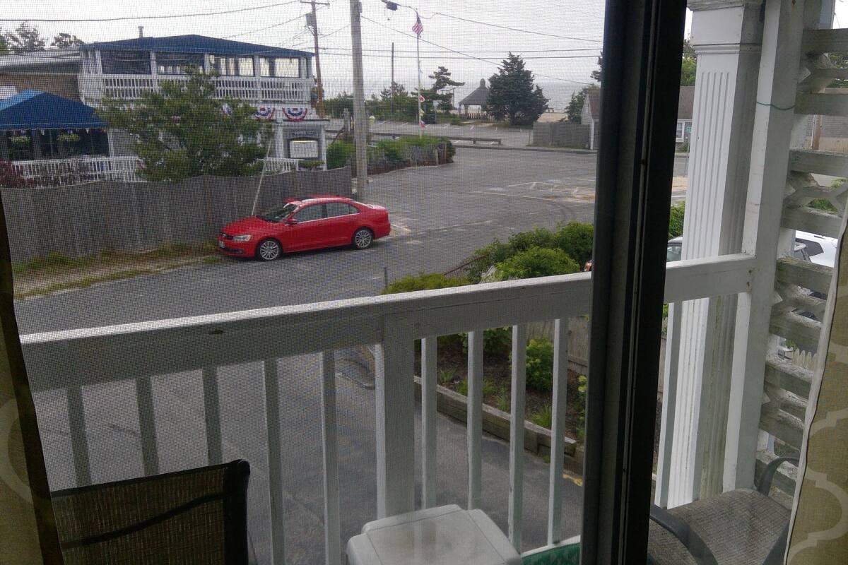 Une vue depuis un balcon, avec une voiture rouge dans la rue, une tente bleue et un bâtiment entouré d’une clôture blanche.