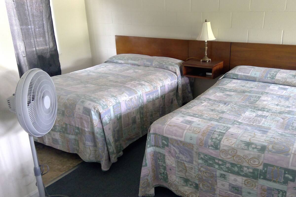 Une chambre d’hôtel avec deux lits simples, une table de chevet avec une lampe, un ventilateur sur pied et une fenêtre avec un rideau.